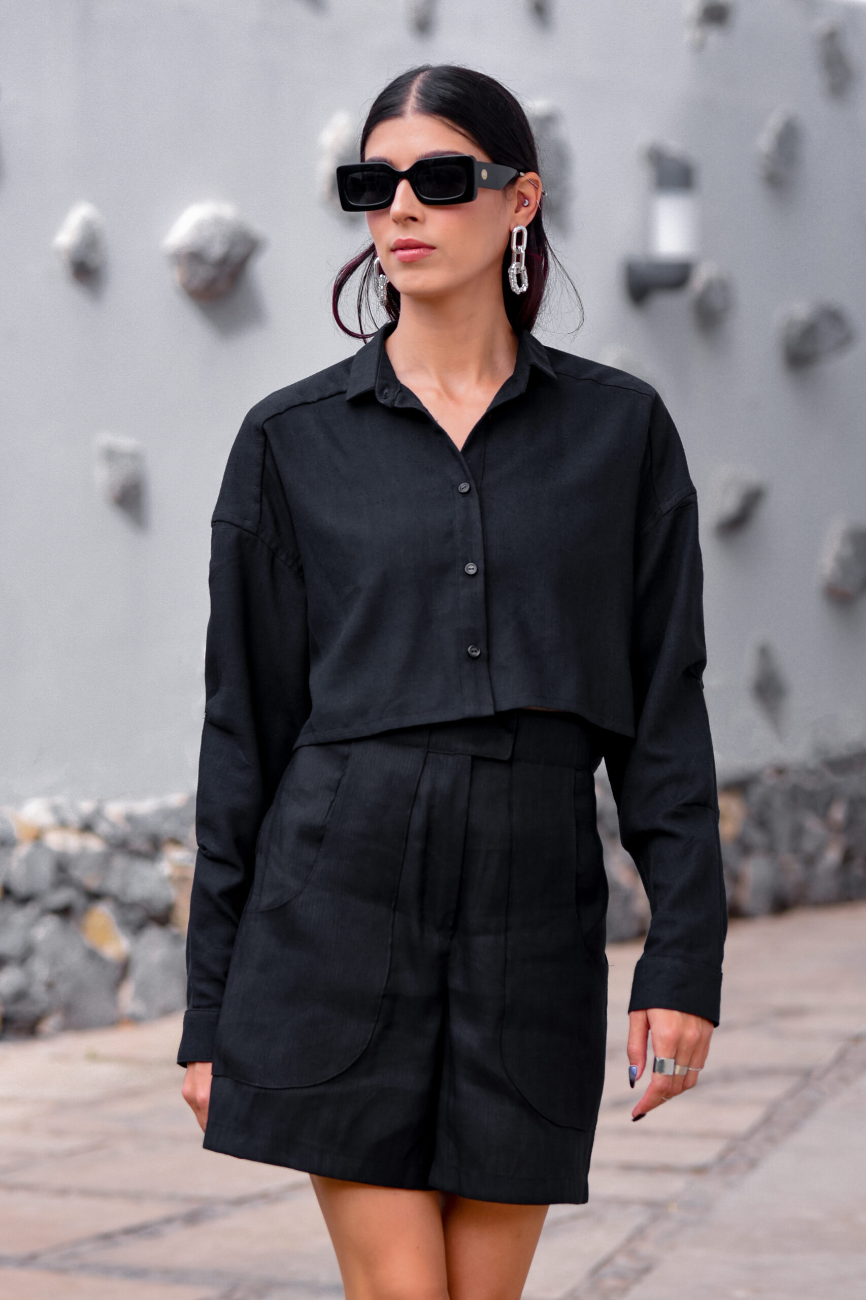 A stylish woman is shown wearing a chic black outfit, consisting of a cropped button-up shirt and high-waisted shorts. She accessorizes with large, statement earrings, dark sunglasses, and a ring on her right hand. Her hair is tied back, and she stands against a minimalist, grey background with a rocky texture. This fashion-forward look is perfect for a modern, urban setting, showcasing contemporary style and elegance.